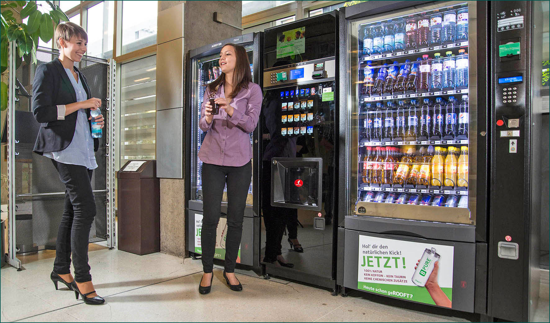 Two women standing near modern vending machines in an office building, enjoying drinks from a vending services dallas